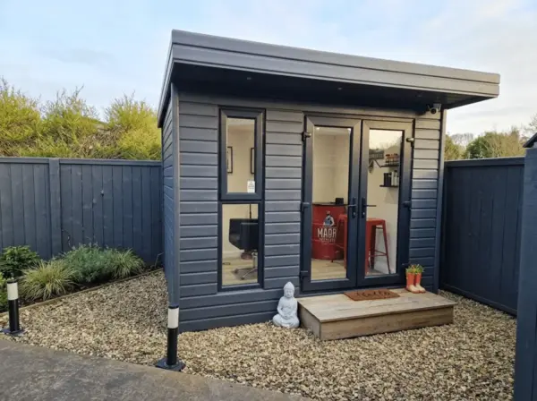 Exterior of The Barbers Den barber shop in Millisle, a modern dark grey timber cabin with glass doors, small porch and landscaped gravel entrance